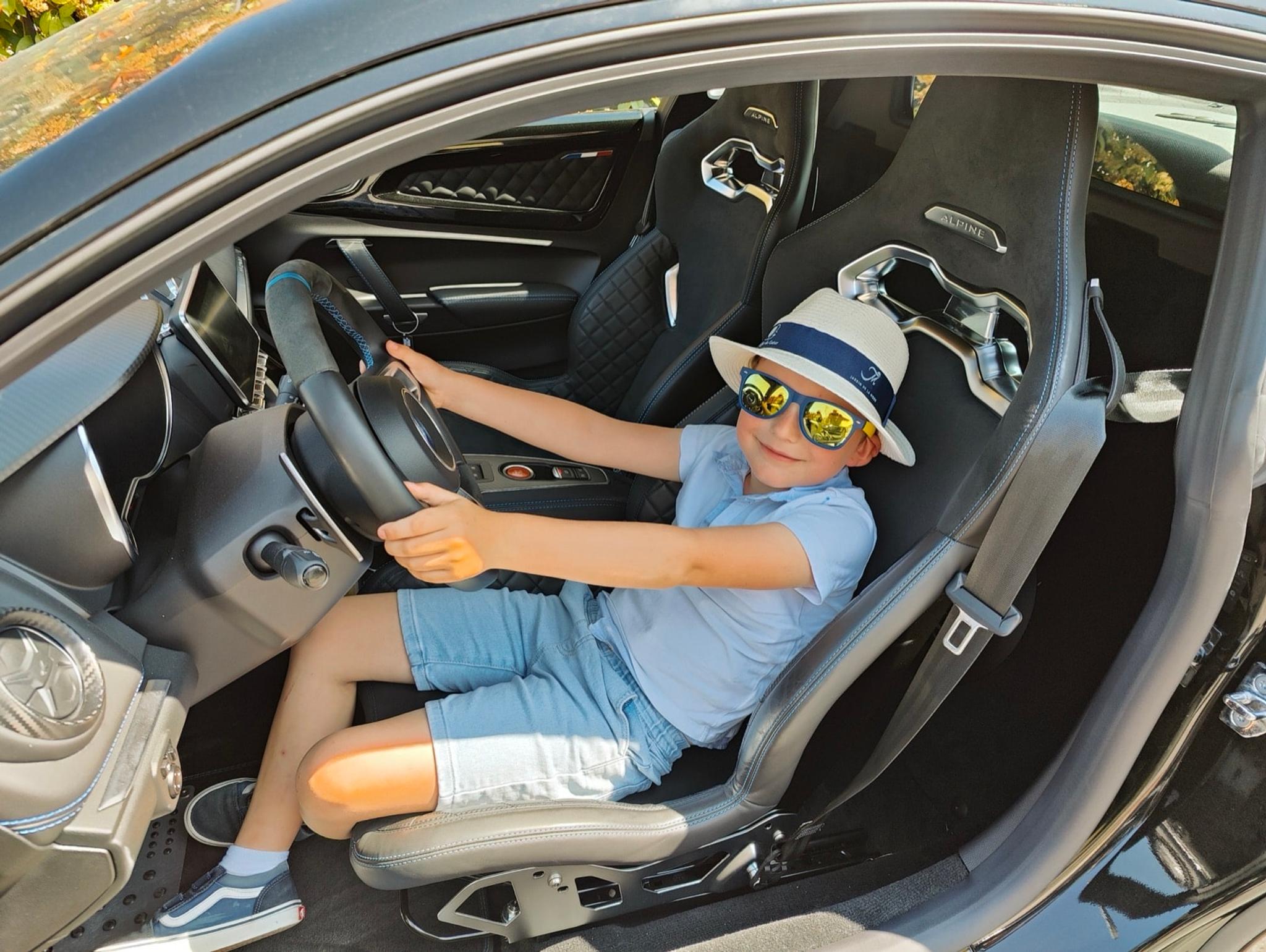 Un enfant en chapeau de paille et lunettes de soleil, tout sourire au volant d'une Alpine A110
